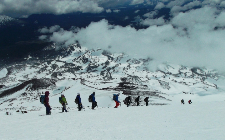 A line of people wearing snow gear, carrying backpacks and using trekking poles, hike through the snow of a mountainous landscape.